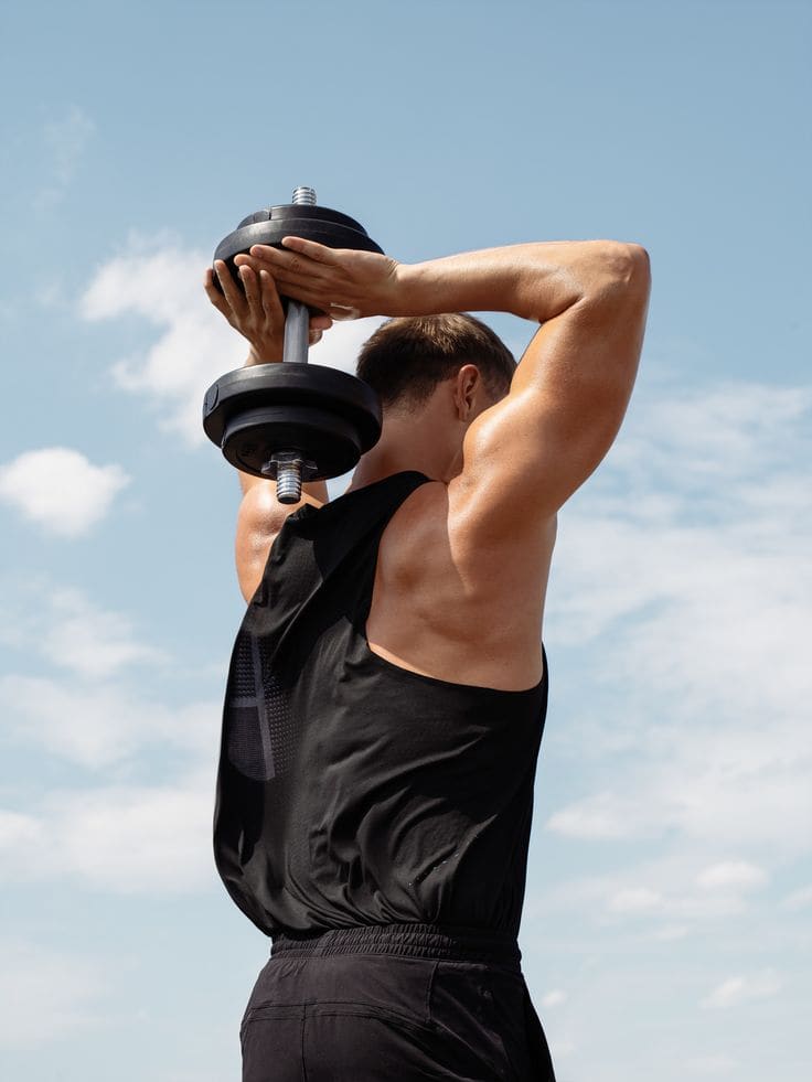 A man pressing a dumbbell overhead against a clear blue sky