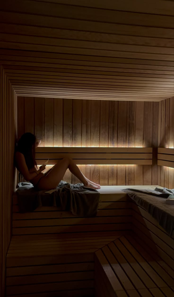 A woman resting on a wooden bench inside a dim, warmly lit sauna