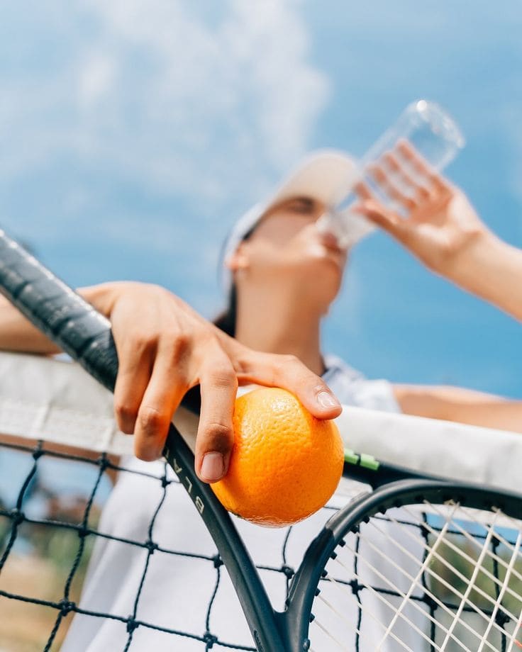 A woman drinking water courtside after a game, holding an orange